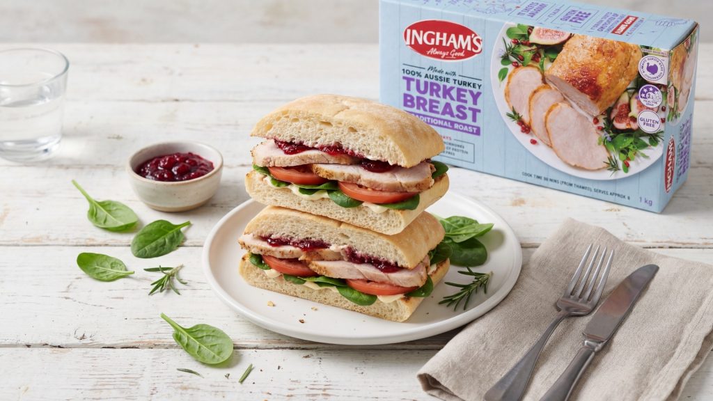 Turkey roll sandwich filled with sliced roast turkey, baby spinach, tomato and cranberry sauce on a white plate, with an Ingham's Turkey Breast Traditional Roast pack in the background.